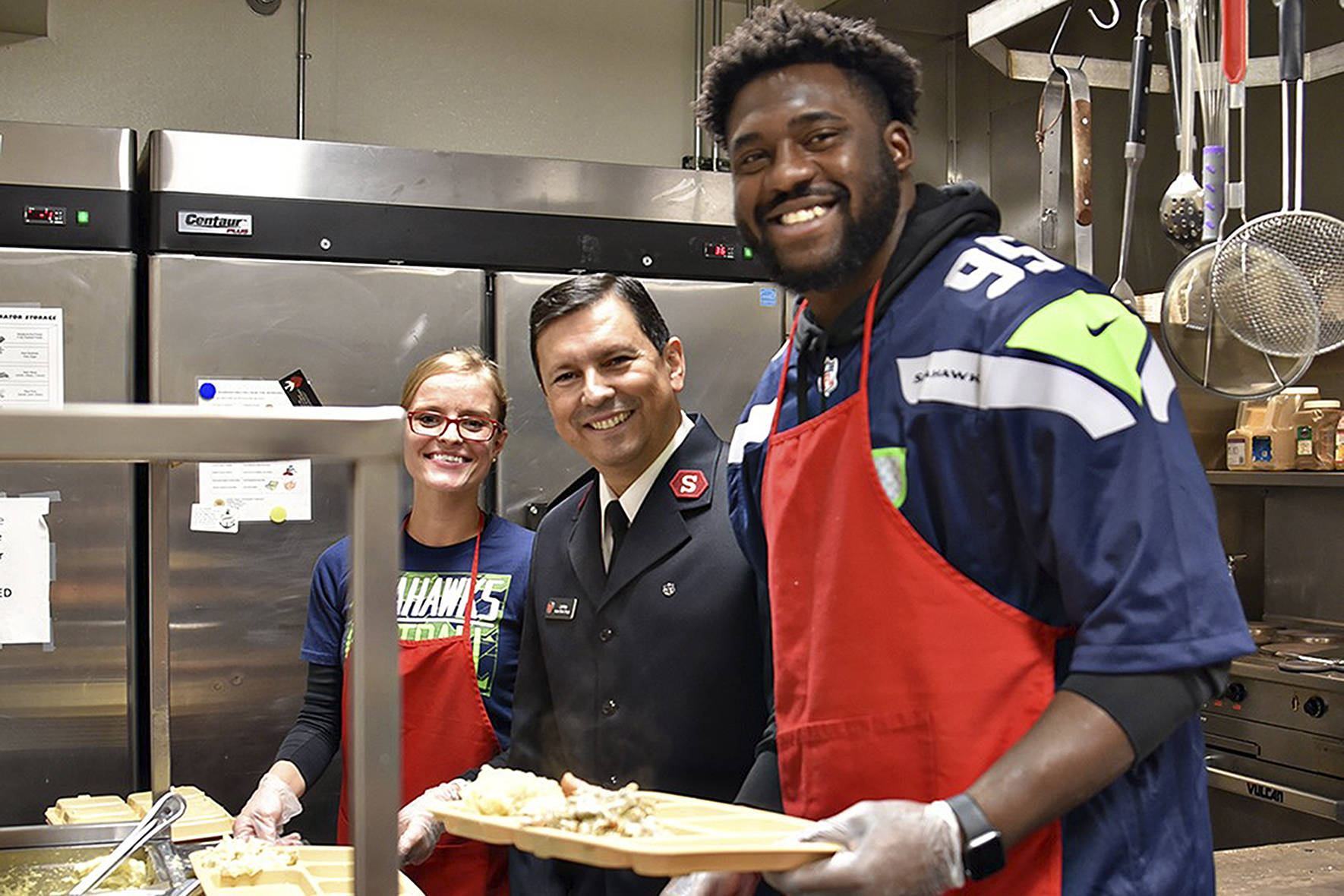 Photo by Danny Rodgers for The Salvation Army.                                Seattle Seahawks Defensive End L.J. Collier volunteers at The Salvation Army community meal on Wednesday, Nov. 26, helping serve a traditional Thanksgiving meal. The Salvation Army serves a free meal to the public, Monday through Thursday, 5:15 to 6:30 p.m. Also pictured are Captain Isaias Braga, director of The Salvation Army in Renton, and Larissa Braga, volunteer.