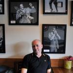 Mick Flynn sits underneath his Blues Greats acrylic series at Liberty Cafe. Photo by Leah Abraham
